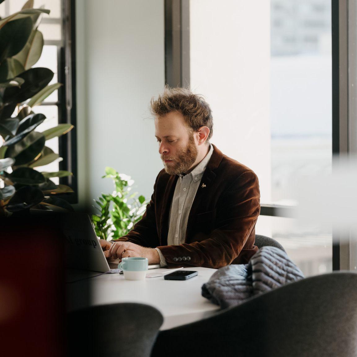 A person in a bright office with big windows is seated and working on a laptop